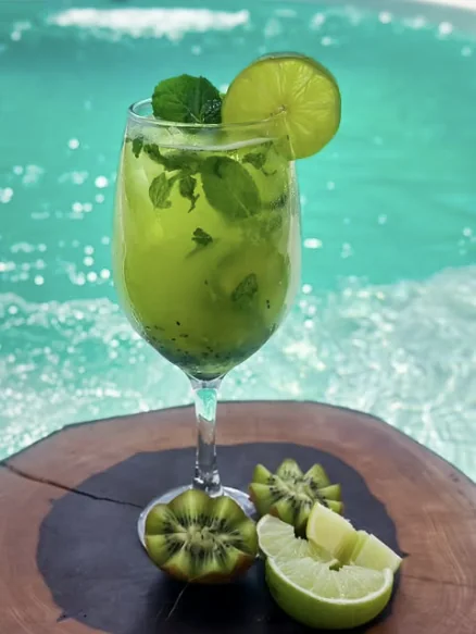Caipirinha de kiwi com hortelã e rodela de limão, ao lado de kiwis cortados.