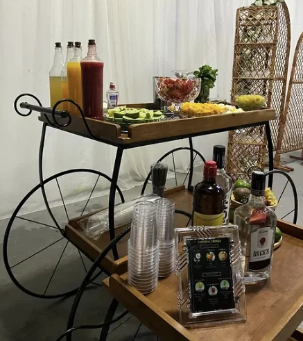 Carrinho de bar de ferro e madeira com garrafas, frutas, copos e carta de drinks.