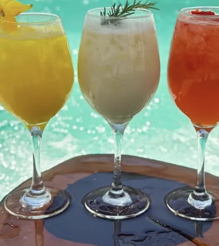 Três drinks em taças: um amarelo de maracujá, um cremoso com alecrim e um vermelho de frutas, à beira da piscina.